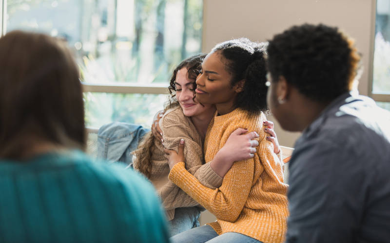A picture of a group therapy session of teens where two teens are hugging each other after experiencing a mental health crisis.